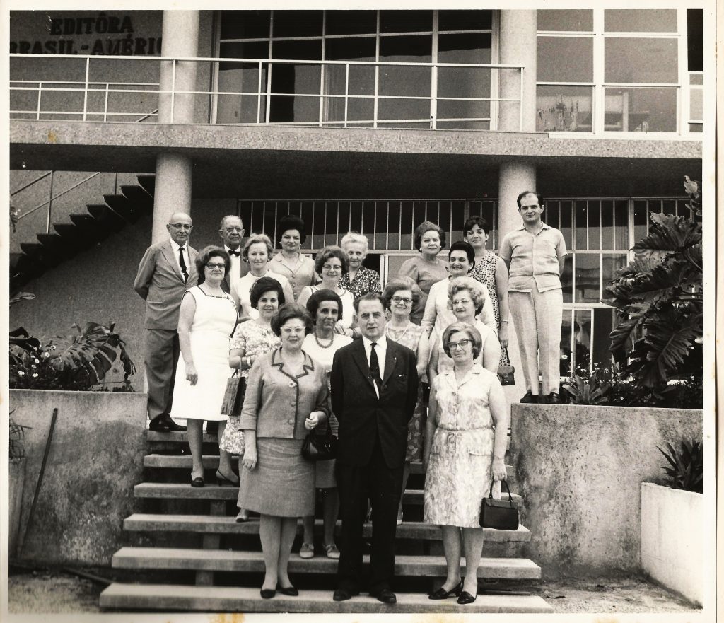A foto está sob um fundo branco, uma foto antiga em preta e branco, possui um grupo de 18 pessoas reunidas olhando para frente e para câmera sorrindo para o fotógrafo, as pessoas estão distribuídas nos degraus de uma escada com algumas pessoas em cada degrau. Ao centro com seu terno preto está o professor, Malba Tahan, com boa postura com as mãos cruzadas atrás do corpo. Ao seu redor, contrastando com a roupa preta do professor, um conjunto, em sua maioria de mulheres entre 30 e 60 anos, usando roupas claras, finamente vestidas, no último degrau existem 2 senhores de terno cinza na ponta esquerda, e um rapaz de roupa branca na ponta direita. O grupo se reúne na entrada de uma construção, a Editora Brasil-América, estão numa escada ladeada por um jardim de plantas com grandes folhas. Ao fundo um par de pilastras cilíndricas que vão do chão, passam a laje do piso do segundo andar e seguem em direção ao teto, no primeiro andar mais ao fundo ao lado direito um portão de painéis de vidro com molduras em alumínio e ao lado esquerdo uma escada que leva ao segundo piso. No segundo piso existe em grandes letras parafusadas na parede, os dizeres "Editora Brasil-América" no alto à esquerda da parede, e à direita uma fachada de com portas em painéis de vidro moldurados com alumínio, sendo protegidos na frente, no primeiro plano, por uma grade baixa de alumínio.